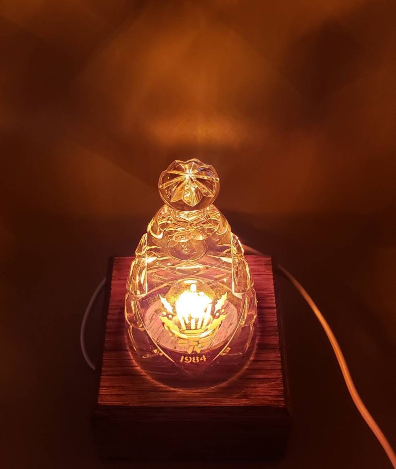 May include: A clear glass lamp base with a crown design and the year 1984 etched on it. The lamp base is sitting on a wooden base.