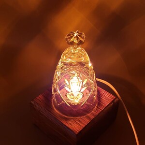 May include: A small, clear glass lamp with a crystal-like design sits on a wooden base. The lamp has a small, round bulb and a decorative design on the front. The lamp is lit and casting a warm glow.