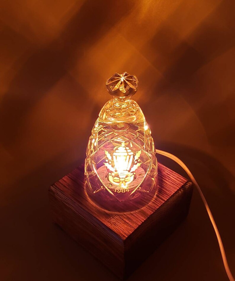 May include: A small, clear glass lamp with a crystal-like design sits on a wooden base. The lamp has a small, round bulb and a decorative design on the front. The lamp is lit and casting a warm glow.