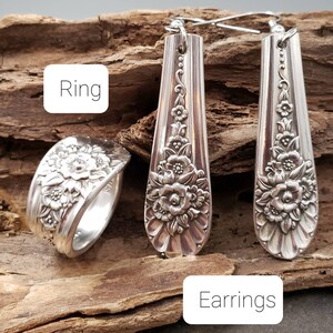 May include: A set of silver earrings and a ring made from vintage silverware. The earrings are long and dangle, with a floral design. The ring is wide and has a floral design.
