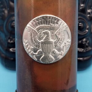 May include: A copper and brass container with a silver-colored eagle emblem on the front. The eagle is surrounded by a circle with the words "E Pluribus Unum" and "One Cent" inscribed on it. The container has a gold-colored band around the top and bottom.