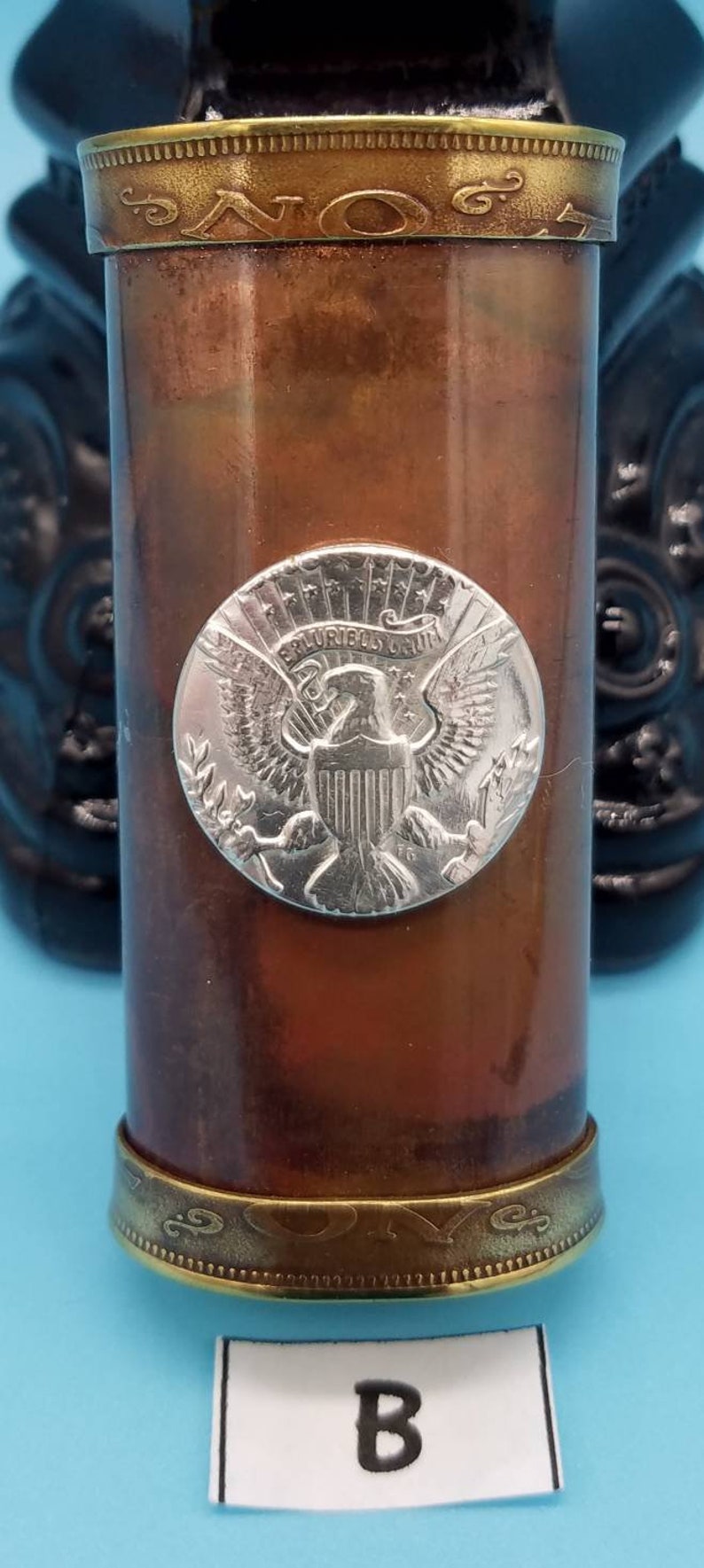 May include: A copper and brass container with a silver-colored eagle emblem on the front. The eagle is surrounded by a circle with the words "E Pluribus Unum" and "One Cent" inscribed on it. The container has a gold-colored band around the top and bottom.