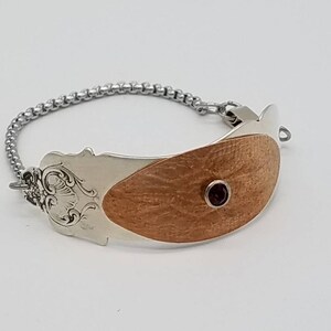 May include: A silver bracelet with a wooden inlay and a small red gemstone. The bracelet is made from a repurposed spoon.