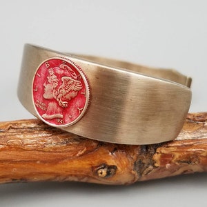 May include: A silver cuff bracelet with a red 1941 Mercury dime embedded in the metal. The coin features the profile of Liberty with wings.