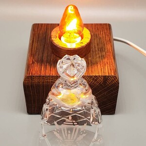 May include: A small, clear glass lampshade with a decorative etched design sits on a wooden base with a lit light bulb inside. The lampshade has a clear glass top and a decorative etched design of a lantern with holly leaves.