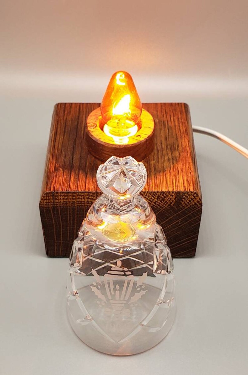 May include: A small, clear glass lampshade with a decorative etched design sits on a wooden base with a lit light bulb inside. The lampshade has a clear glass top and a decorative etched design of a lantern with holly leaves.