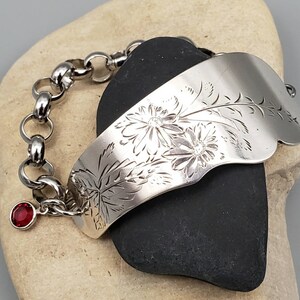 May include: A silver bracelet with a floral design and a red gemstone charm. The bracelet is made from a repurposed spoon and features a chain link closure.