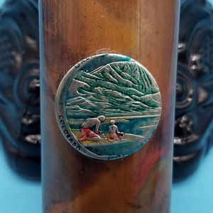 May include: A copper-colored cylindrical container with a brass top and bottom. The container has a round, silver-colored sticker with a green mountain scene and the words "Colorado" and "U.S.A." on it.