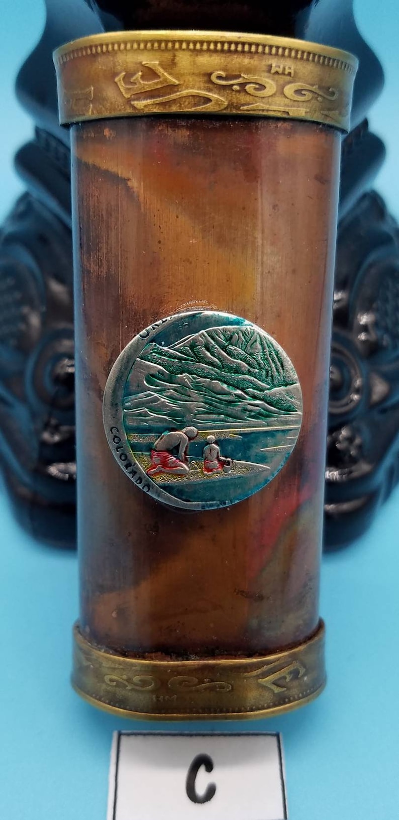 May include: A copper-colored cylindrical container with a brass top and bottom. The container has a round, silver-colored sticker with a green mountain scene and the words "Colorado" and "U.S.A." on it.
