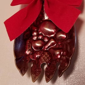 May include: A red velvet bow with a gold accent is attached to a red metal ornament shaped like a fork with a fruit design. The ornament is labeled with the letter "A".