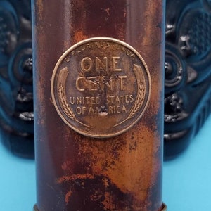 May include: A copper-colored cylinder with a gold top and bottom. The cylinder has a penny embedded in the side, with the inscription "ONE CENT, UNITED STATES OF AMERICA".