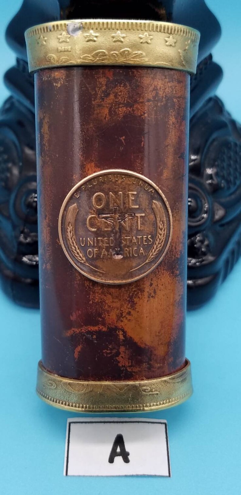 May include: A copper-colored cylinder with a gold top and bottom. The cylinder has a penny embedded in the side, with the inscription "ONE CENT, UNITED STATES OF AMERICA".