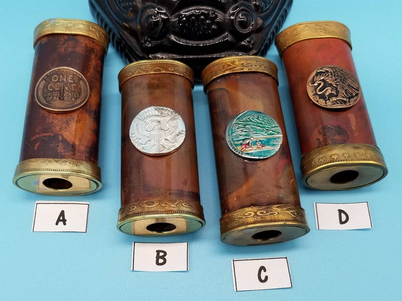 May include: Four decorative wooden tubes with different coin designs on the top. The tubes are brown with gold trim and have a hole at the bottom. The coins are a US penny, a US silver dollar, a coin with a mountain scene, and a coin with an eagle design.