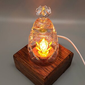 May include: A clear glass lamp with a gold-colored design and the year 1984 inscribed on the bottom. The lamp is lit and sits on a wooden base.