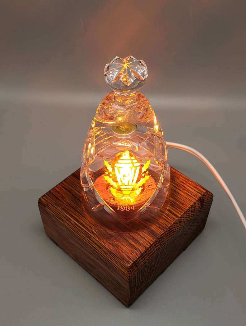 May include: A clear glass lamp with a gold-colored design and the year 1984 inscribed on the bottom. The lamp is lit and sits on a wooden base.