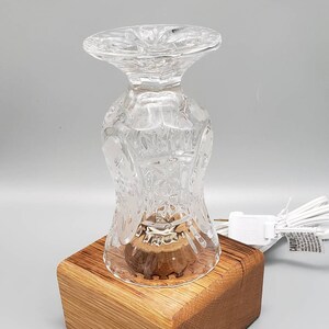 Puede incluir: Una base de l&aacute;mpara de vidrio transparente con un dise&ntilde;o de vidrio cortado se asienta sobre una base de madera. La l&aacute;mpara est&aacute; encendida y tiene un cable con un enchufe.