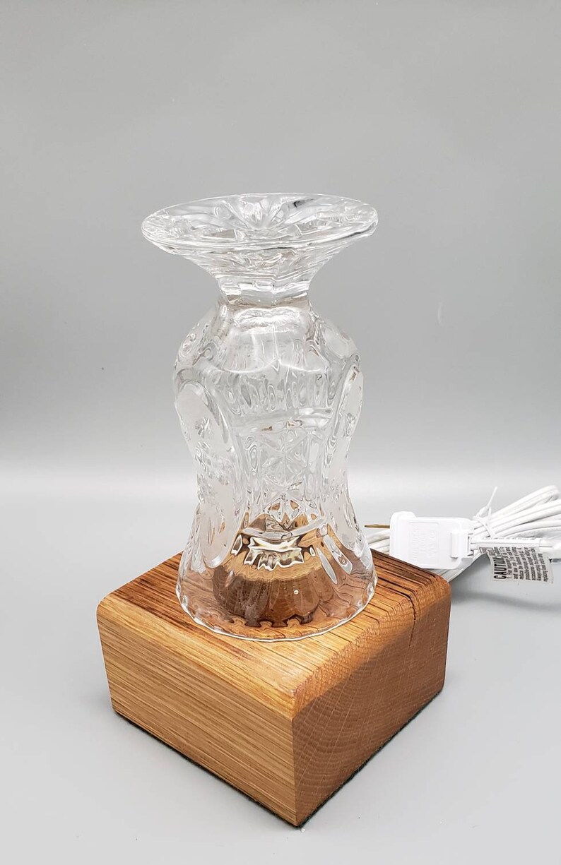 Puede incluir: Una base de l&aacute;mpara de vidrio transparente con un dise&ntilde;o de vidrio cortado se asienta sobre una base de madera. La l&aacute;mpara est&aacute; encendida y tiene un cable con un enchufe.