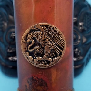 May include: A copper and brass shot glass with a gold rim and a decorative eagle design. The eagle is holding a snake in its talons. The glass has a star pattern around the top and bottom.