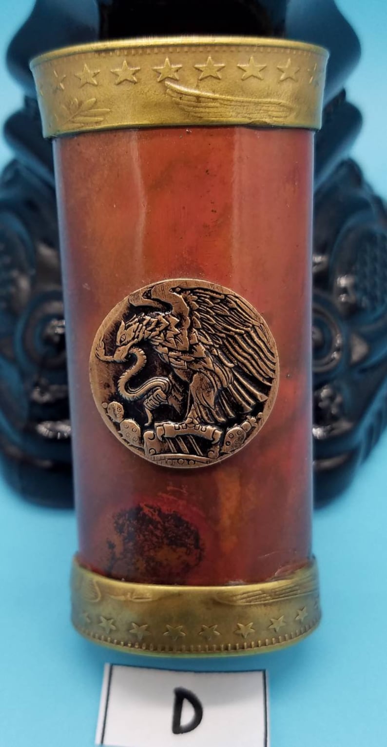 May include: A copper and brass shot glass with a gold rim and a decorative eagle design. The eagle is holding a snake in its talons. The glass has a star pattern around the top and bottom.