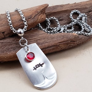 May include: A silver necklace with a silver dog tag pendant. The pendant has a red gemstone and the inscription "424" on it.