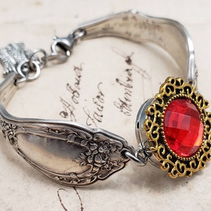 May include: A silver spoon bracelet with a red rhinestone charm. The bracelet is made from a vintage spoon and has a floral design.