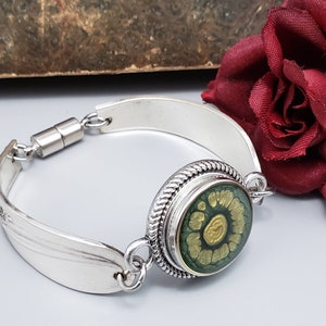 May include: A silver spoon bracelet with a magnetic clasp and a green and gold round charm. The charm has a swirling pattern.