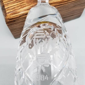May include: A clear glass bell with a diamond pattern and etched design. The bell has a gold rim and the number 9834 is etched on the bottom.