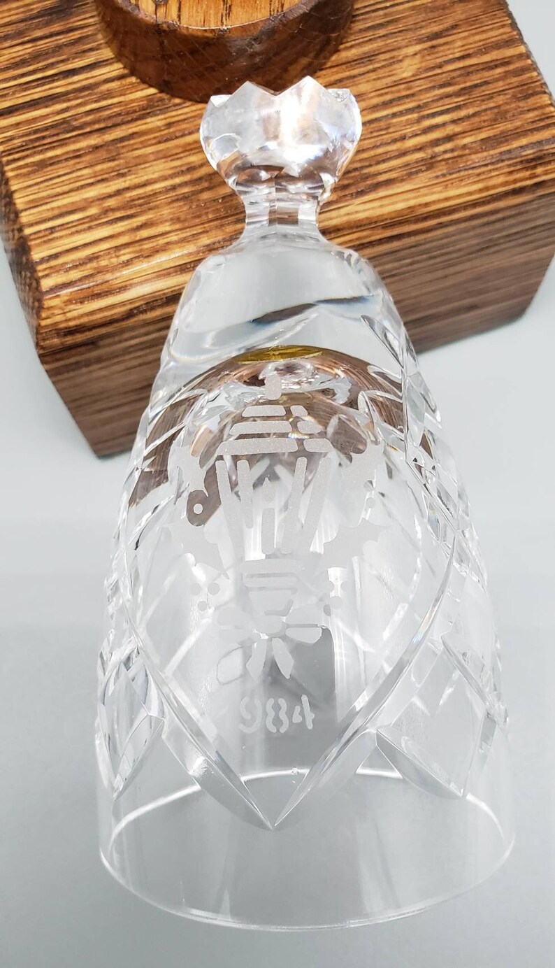 May include: A clear glass bell with a diamond pattern and etched design. The bell has a gold rim and the number 9834 is etched on the bottom.
