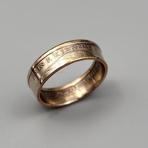 May include: A gold-toned ring made from a half-dollar coin featuring the inscription "JOHN F. KENNEDY" and "UNITED STATES OF AMERICA".