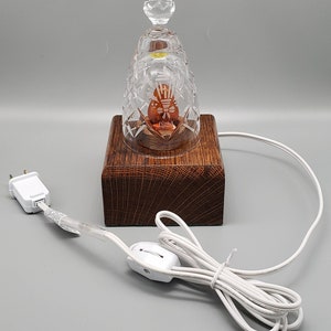 May include: A clear glass bell with a crystal design and a small light bulb inside. The bell is sitting on a wooden base with a cord and switch.