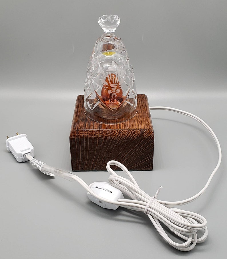 May include: A clear glass bell with a crystal design and a small light bulb inside. The bell is sitting on a wooden base with a cord and switch.