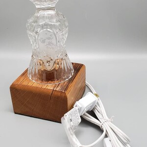 Puede incluir: Una base de l&aacute;mpara de vidrio transparente con un patr&oacute;n decorativo se asienta sobre una base de madera con un cable el&eacute;ctrico blanco. La l&aacute;mpara est&aacute; enchufada a una toma de corriente.