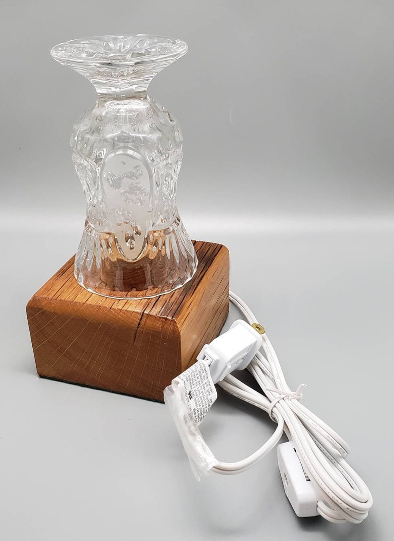 Puede incluir: Una base de l&aacute;mpara de vidrio transparente con un patr&oacute;n decorativo se asienta sobre una base de madera con un cable el&eacute;ctrico blanco. La l&aacute;mpara est&aacute; enchufada a una toma de corriente.