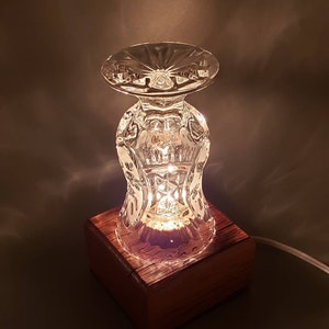 Puede incluir: Una base de l&aacute;mpara de vidrio hecha de un vaso de cristal vintage, con una base de madera. La l&aacute;mpara est&aacute; encendida y proyecta un brillo c&aacute;lido.