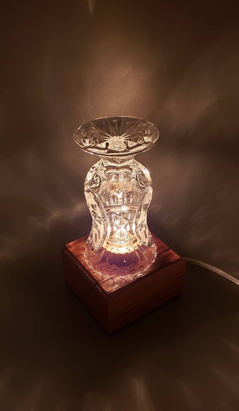 Puede incluir: Una base de l&aacute;mpara de vidrio hecha de un vaso de cristal vintage, con una base de madera. La l&aacute;mpara est&aacute; encendida y proyecta un brillo c&aacute;lido.