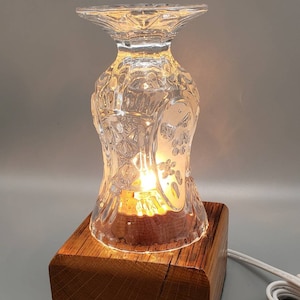 Puede incluir: Una peque&ntilde;a l&aacute;mpara de mesa hecha a partir de una copa de vidrio reutilizada con una bombilla en su interior. La copa es transparente y tiene un dise&ntilde;o de vidrio cortado. La base de la l&aacute;mpara es un trozo de madera cuadrado.