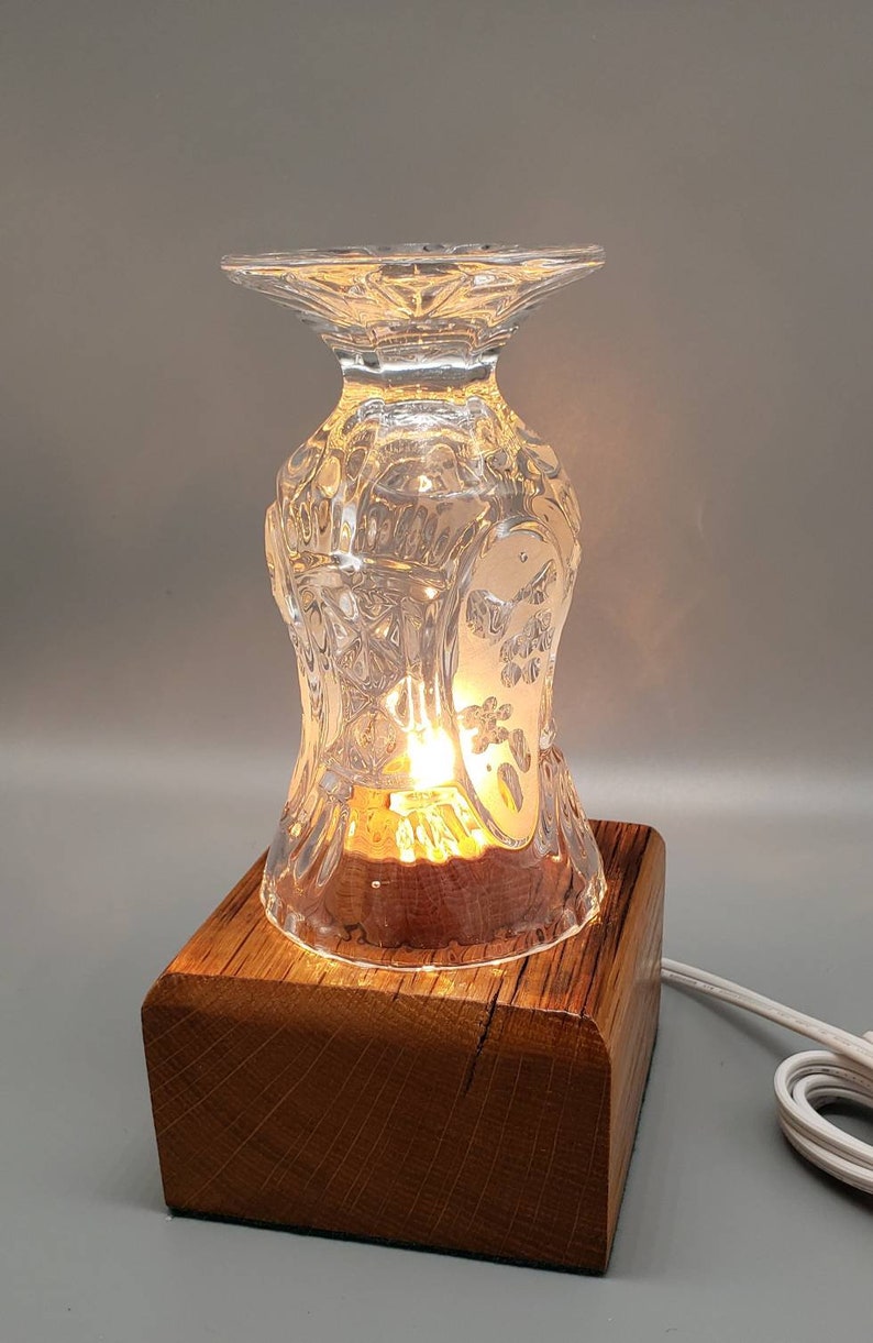 Puede incluir: Una peque&ntilde;a l&aacute;mpara de mesa hecha a partir de una copa de vidrio reutilizada con una bombilla en su interior. La copa es transparente y tiene un dise&ntilde;o de vidrio cortado. La base de la l&aacute;mpara es un trozo de madera cuadrado.