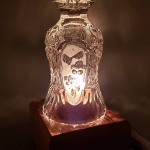 Puede incluir: Una peque&ntilde;a l&aacute;mpara de mesa hecha de una copa de vidrio transparente con un patr&oacute;n decorativo y una base de madera. La l&aacute;mpara est&aacute; encendida y proyecta un brillo c&aacute;lido.