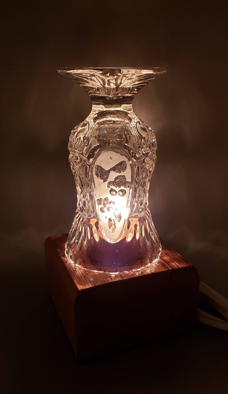 Puede incluir: Una peque&ntilde;a l&aacute;mpara de mesa hecha de una copa de vidrio transparente con un patr&oacute;n decorativo y una base de madera. La l&aacute;mpara est&aacute; encendida y proyecta un brillo c&aacute;lido.