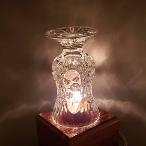 Puede incluir: Una peque&ntilde;a l&aacute;mpara de vidrio transparente con un dise&ntilde;o texturizado se asienta sobre una base de madera. La l&aacute;mpara est&aacute; encendida y emite un brillo c&aacute;lido.