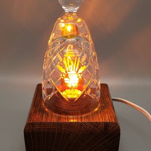 May include: A crystal glass lamp with a wooden base. The lamp is lit and the light shines through the crystal, creating a warm glow. The glass has a decorative design etched into it.