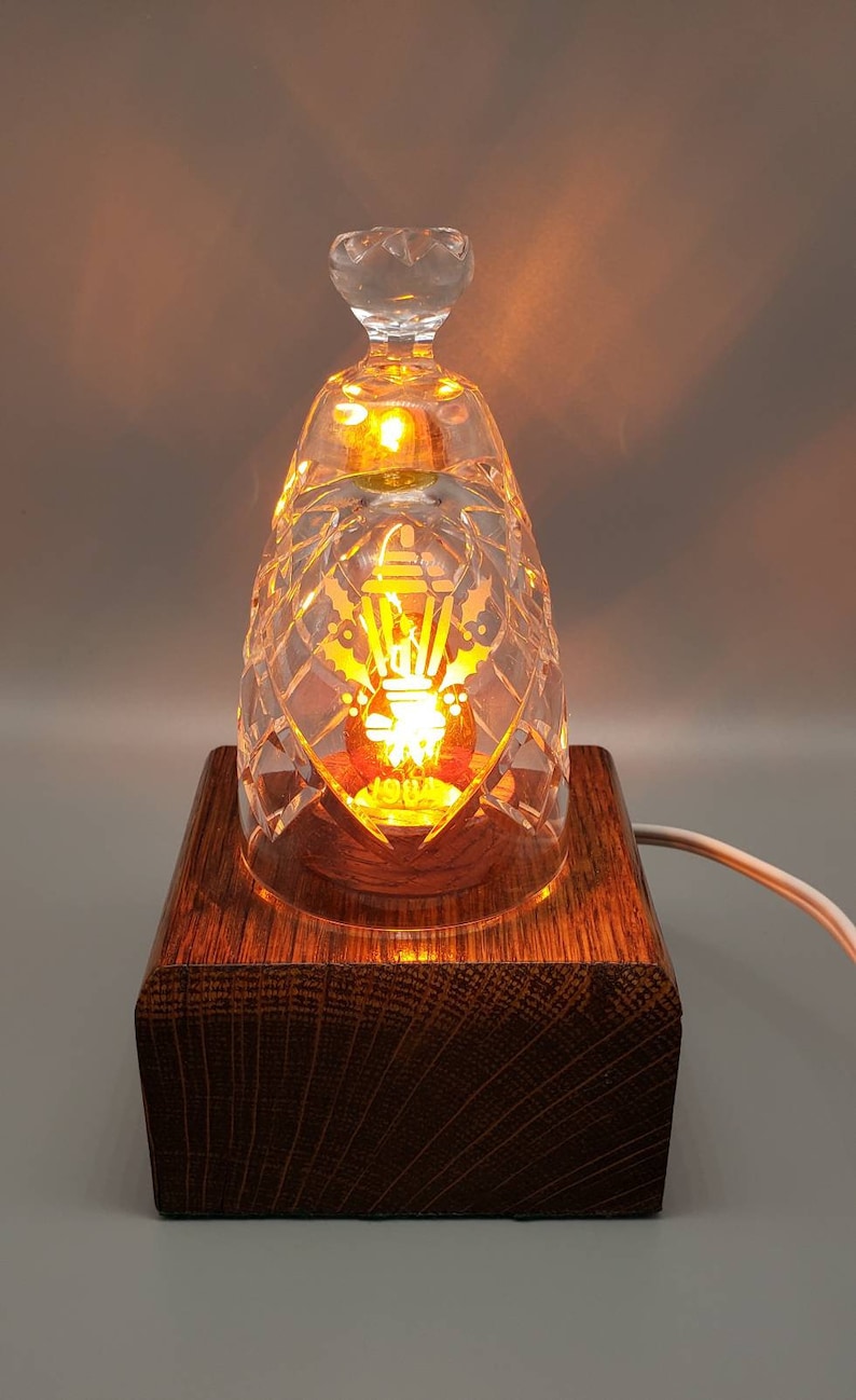 May include: A crystal glass lamp with a wooden base. The lamp is lit and the light shines through the crystal, creating a warm glow. The glass has a decorative design etched into it.