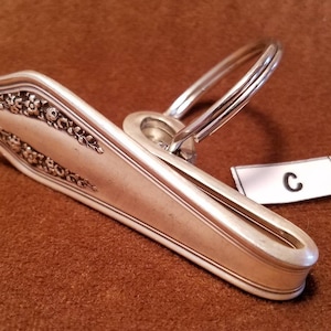 May include: A silver-colored, vintage-style napkin holder with floral detailing and a ring handle. The napkin holder is resting on a brown surface. A small white tag with the letter "C" is attached.