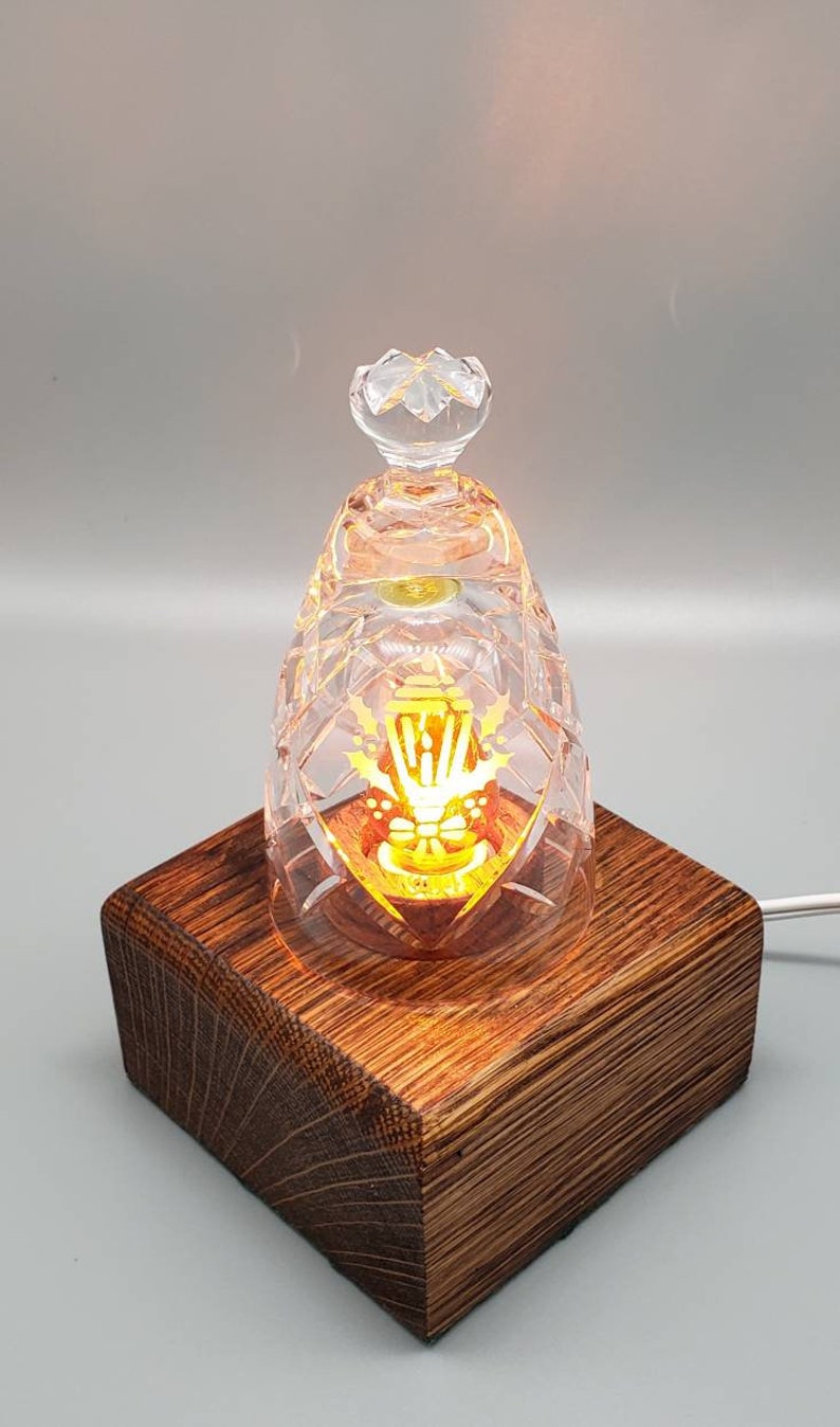 May include: A crystal glass lampshade with a gold design sits on a dark brown wooden base. The lampshade is lit from within, casting a warm glow.