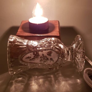 Puede incluir: Un jarr&oacute;n de vidrio transparente con un dise&ntilde;o decorativo se encuentra sobre una base de madera con una vela de t&eacute; encendida en su interior. El jarr&oacute;n est&aacute; inclinado hacia un lado, y la luz de la vela proyecta un c&aacute;lido resplandor sobre la superficie.