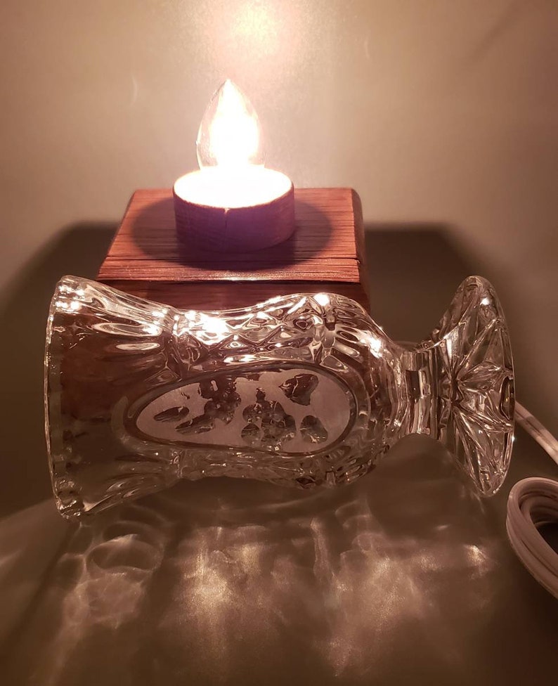 Puede incluir: Un jarr&oacute;n de vidrio transparente con un dise&ntilde;o decorativo se encuentra sobre una base de madera con una vela de t&eacute; encendida en su interior. El jarr&oacute;n est&aacute; inclinado hacia un lado, y la luz de la vela proyecta un c&aacute;lido resplandor sobre la superficie.