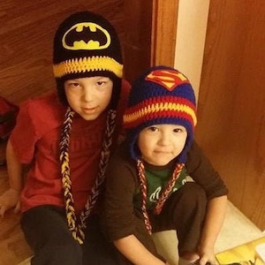 May include: Two children wearing crocheted hats with superhero logos. The boy on the left is wearing a black and yellow Batman hat, and the boy on the right is wearing a red, blue, and yellow Superman hat. Both boys are wearing black pants and sneakers.