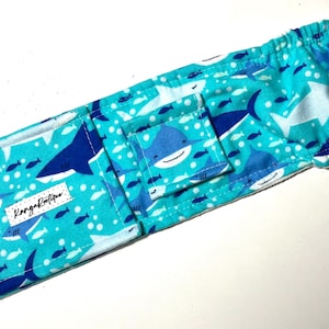 May include: A turquoise blue fabric baby carrier with a white background and a shark print. The carrier has a pocket on the front with a blue shark with a white belly and a white smile. The text "Kangaroo Couture" is printed on the fabric.