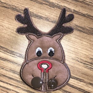 May include: A brown felt reindeer face with a red nose and black eyes. The reindeer has two brown felt antlers. There are two brown buttons on the bottom of the reindeer.