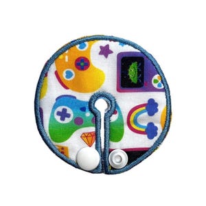 May include: A white, circular fabric item with a blue stitched border, featuring a colorful video game controller pattern. The design includes a rainbow, stars, and a retro video game screen. Two white snaps are attached.